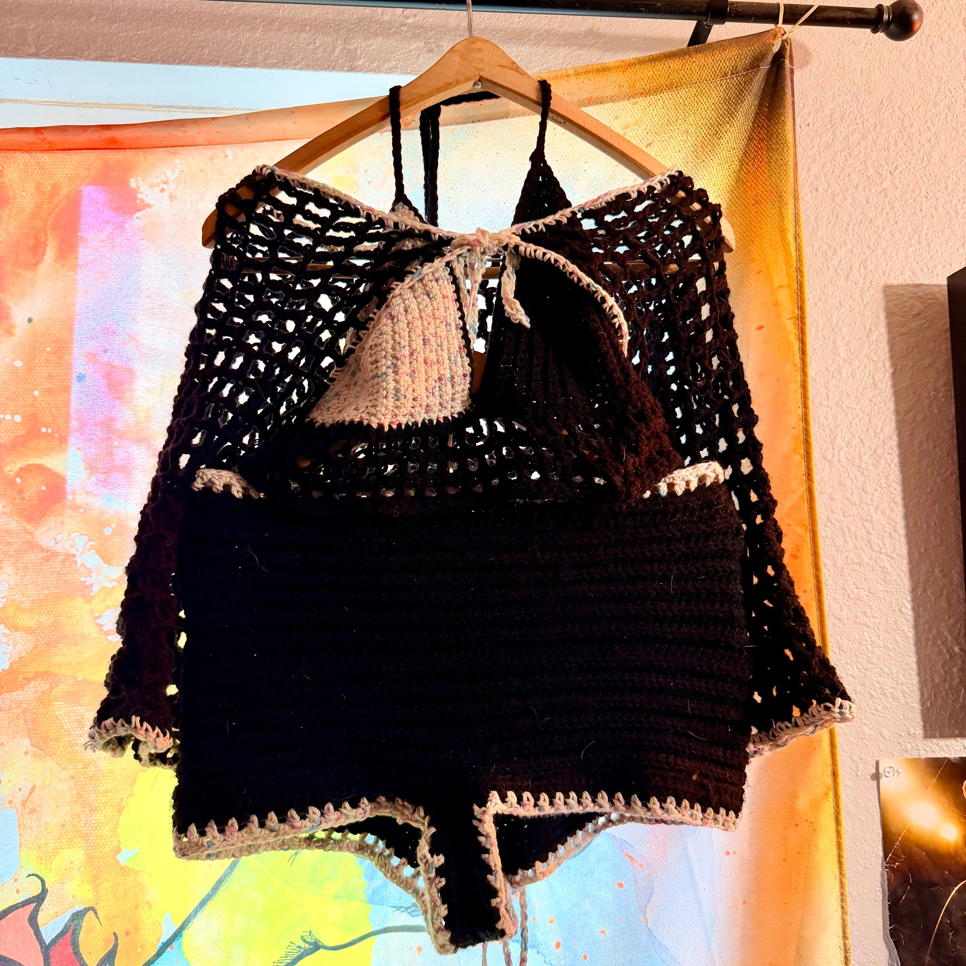 Black crochet top and shorts set on a hanger against a colorful abstract wall.