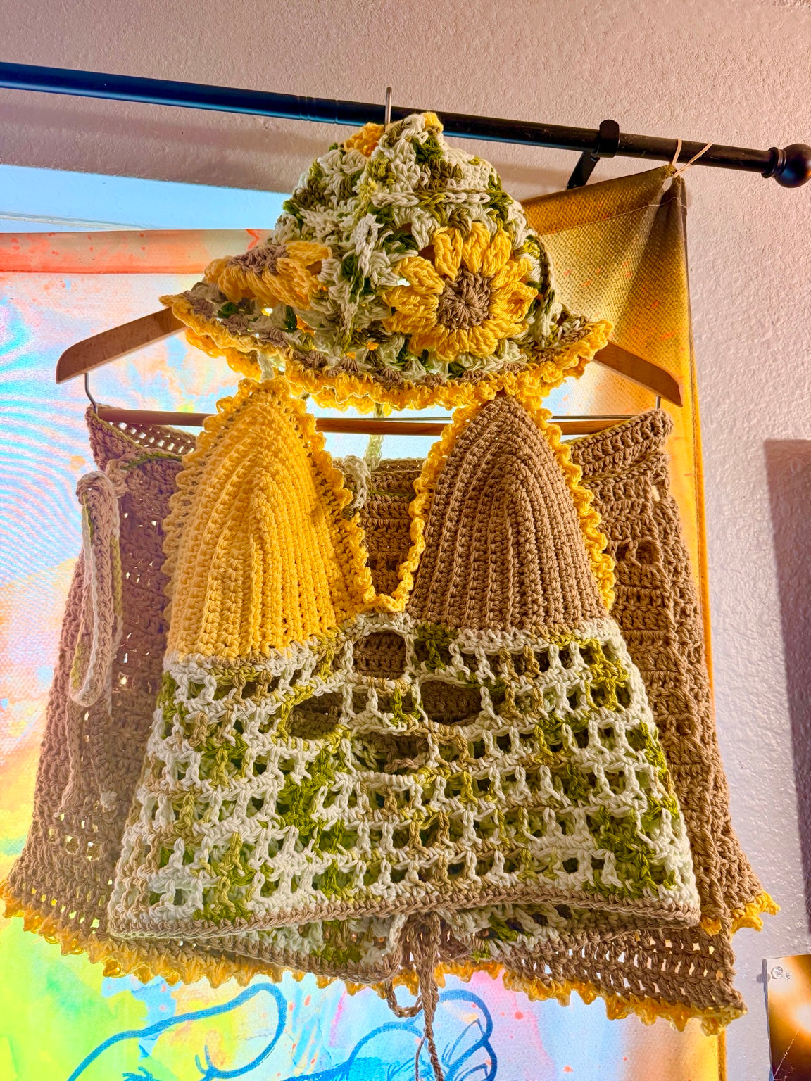 Crocheted festival outfit with a hat and dress on a hanger against a colorful abstract background.